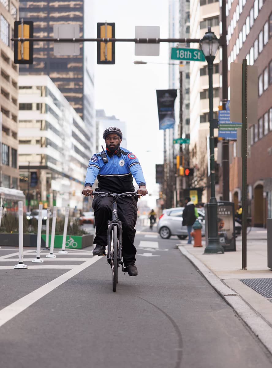 A uniformed cyclist rides confidently down an urban street, lined with tall buildings under a clear sky. The street sign reads 
