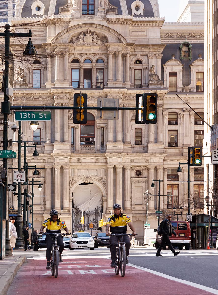 Two cyclists wearing yellow jackets ride on a red bike lane towards a historic, ornate building under a green traffic light on a city street.