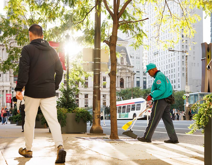 A city worker sweeps the sidewalk near a tree in bright daylight. A person walks by, while urban buildings and a bus are visible in the background.