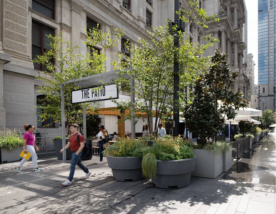 Urban patio scene with people walking past potted greenery. A white sign reads 