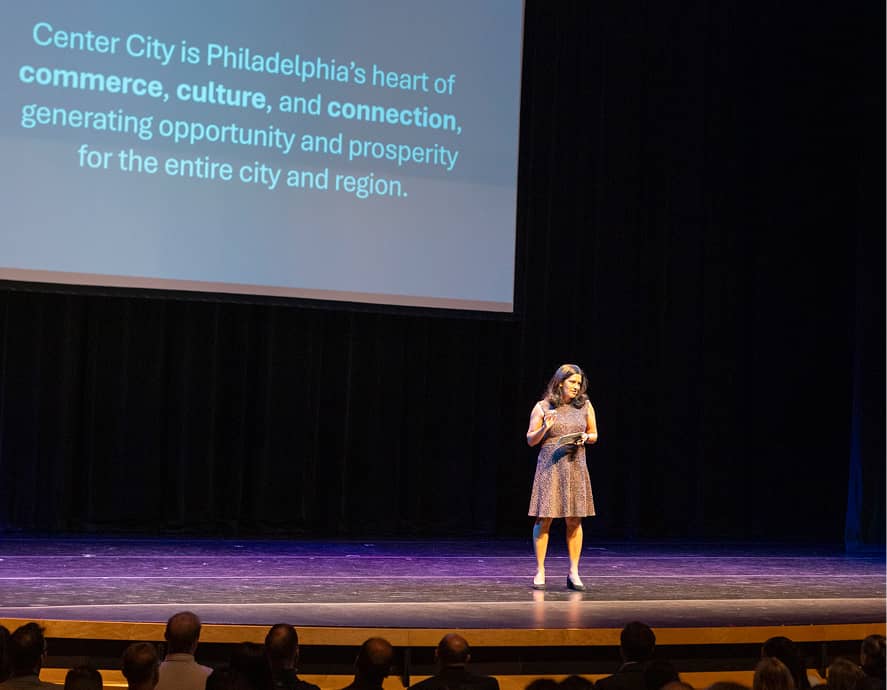A woman stands on an auditorium stage, presenting. Behind her, a slide reads, 