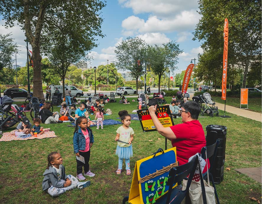A storyteller in a red shirt engages children in a park filled with families and strollers. Bright banners and a clear sky create a cheerful, lively scene.