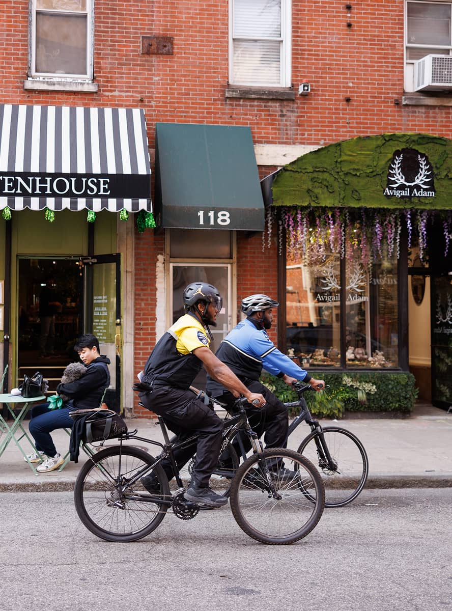 Two cyclists ride past brick buildings with storefronts. A person seated at a table holds a dog, evoking a casual, lively street scene.