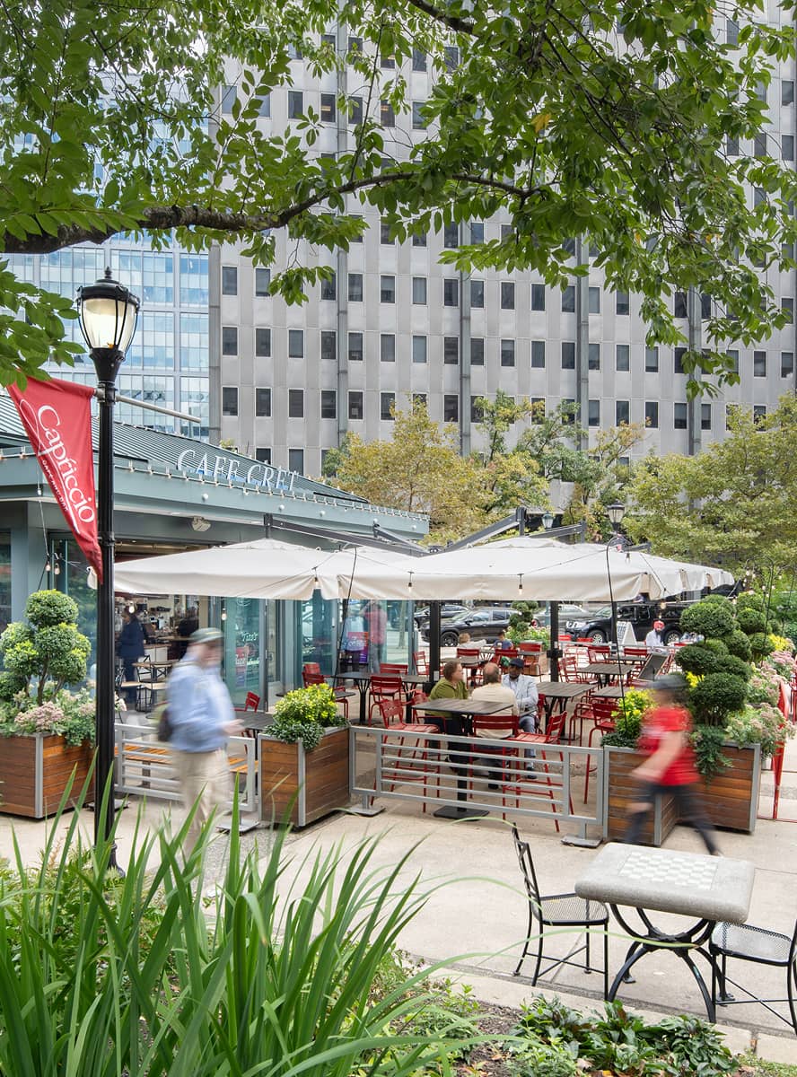 Outdoor cafe with red chairs and white umbrellas on a busy city street. Greenery and trees surround the seating area. People walking and relaxing, creating a lively, inviting atmosphere.