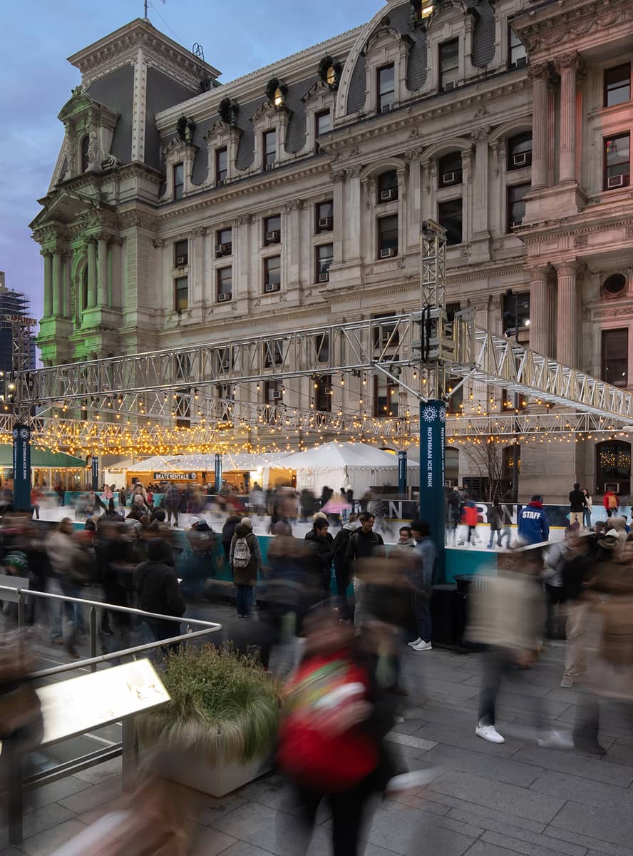 People bustle around a festive outdoor ice skating rink adorned with string lights outside a grand, historic building at dusk, creating a lively atmosphere.