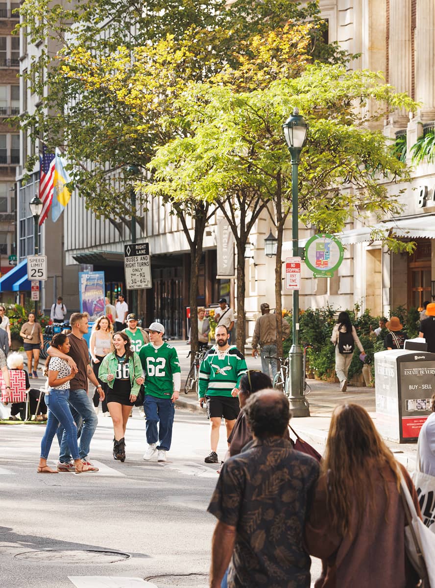 A lively city street scene with people walking past, some in green sports jerseys. Trees and flags line the sidewalk. The atmosphere is casual and bustling.