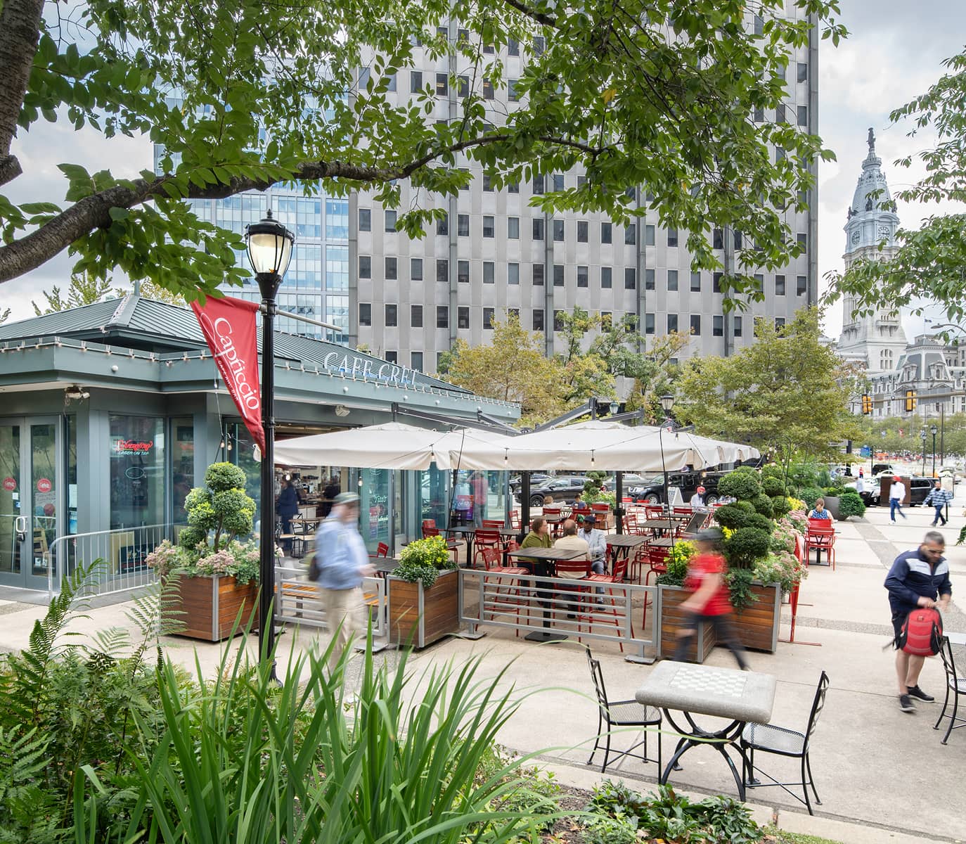 Outdoor cafe with red chairs and large umbrellas on a bustling city sidewalk. People are walking by, and lush greenery surrounds the seating area.