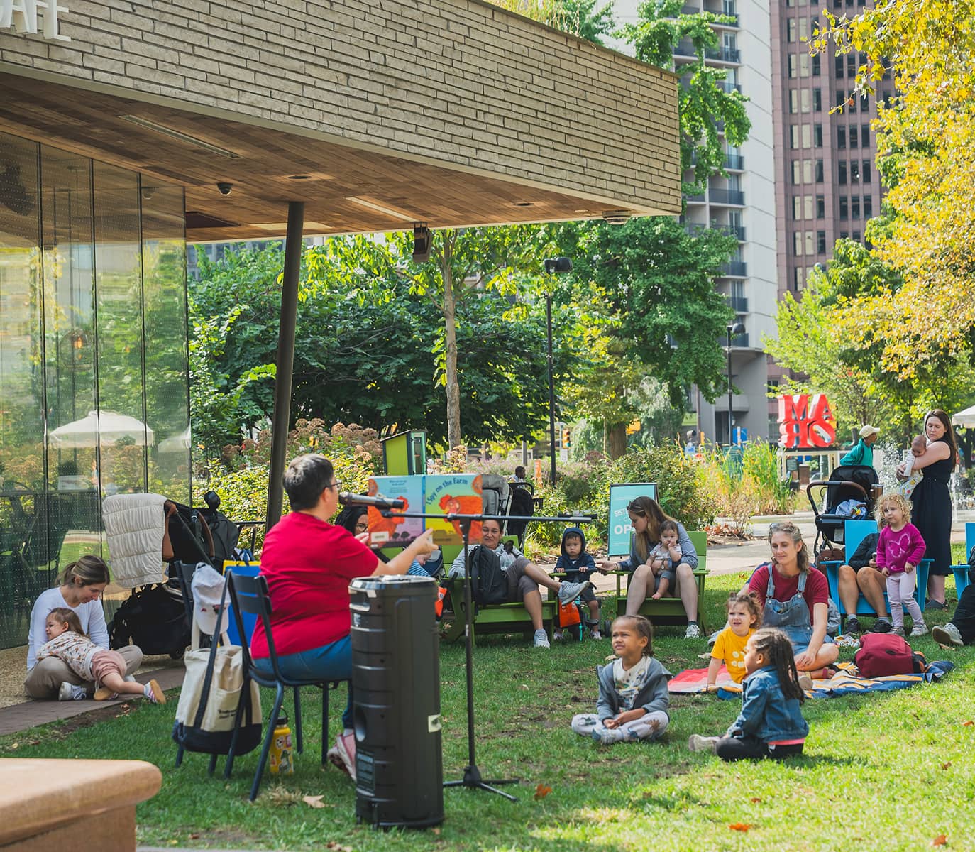 A group of children and adults sit on grass outside a building, engaged in a storytelling session. A storyteller in red holds a book, while kids listen attentively. Trees and city buildings are visible in the background, creating a lively, community-focused atmosphere.