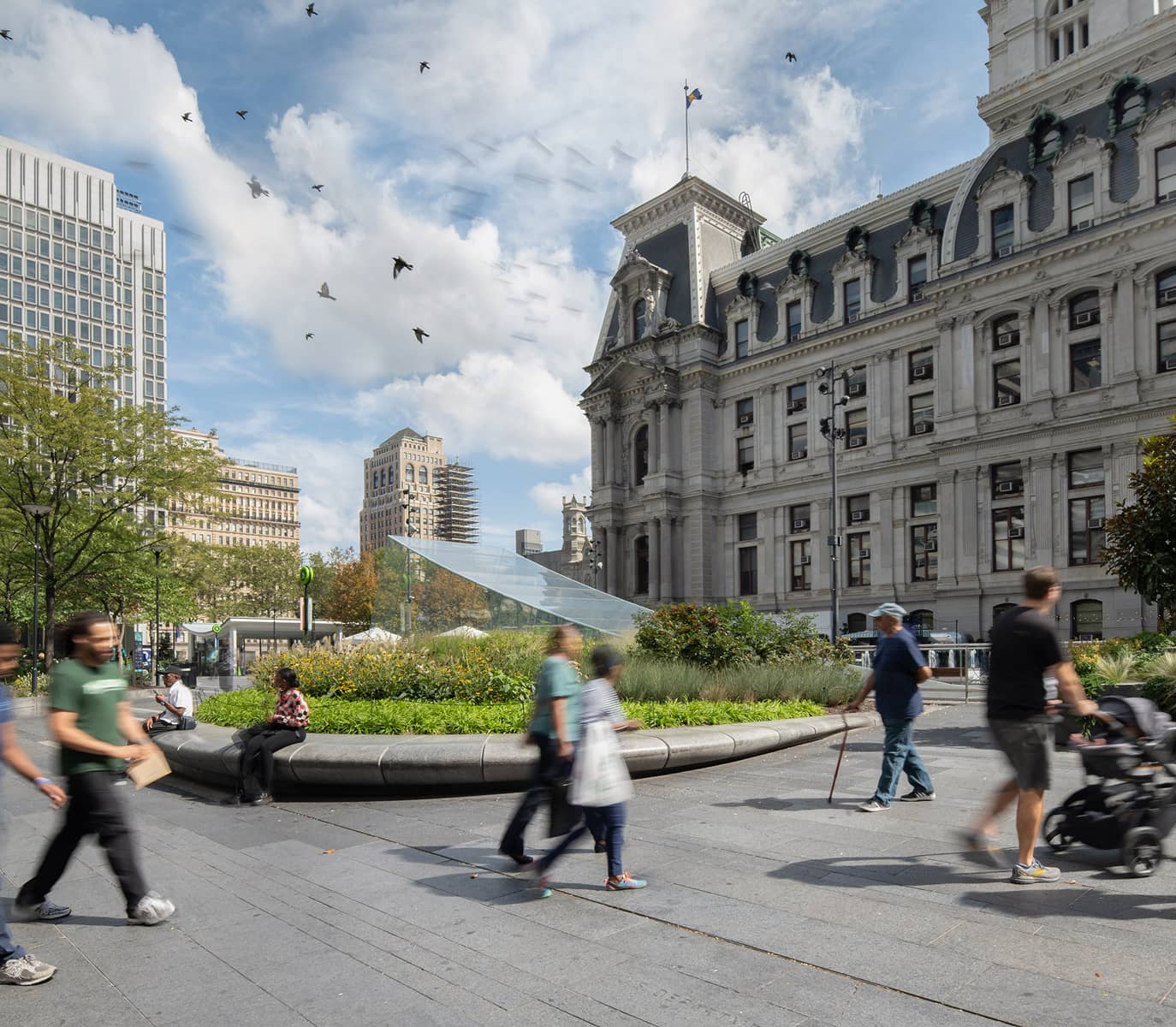 A bustling urban square with people walking, a historic building, lush green landscaping, and birds flying against a bright blue, cloudy sky.