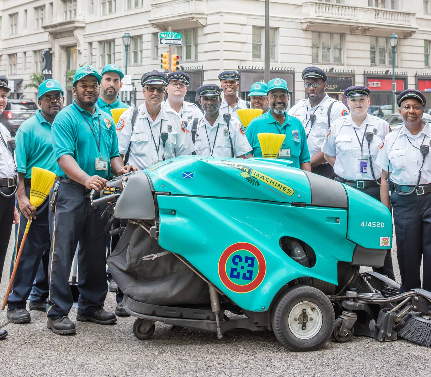 A group of uniformed workers, some in teal shirts and others in white, stand smiling behind a teal street cleaning machine, on a city street.