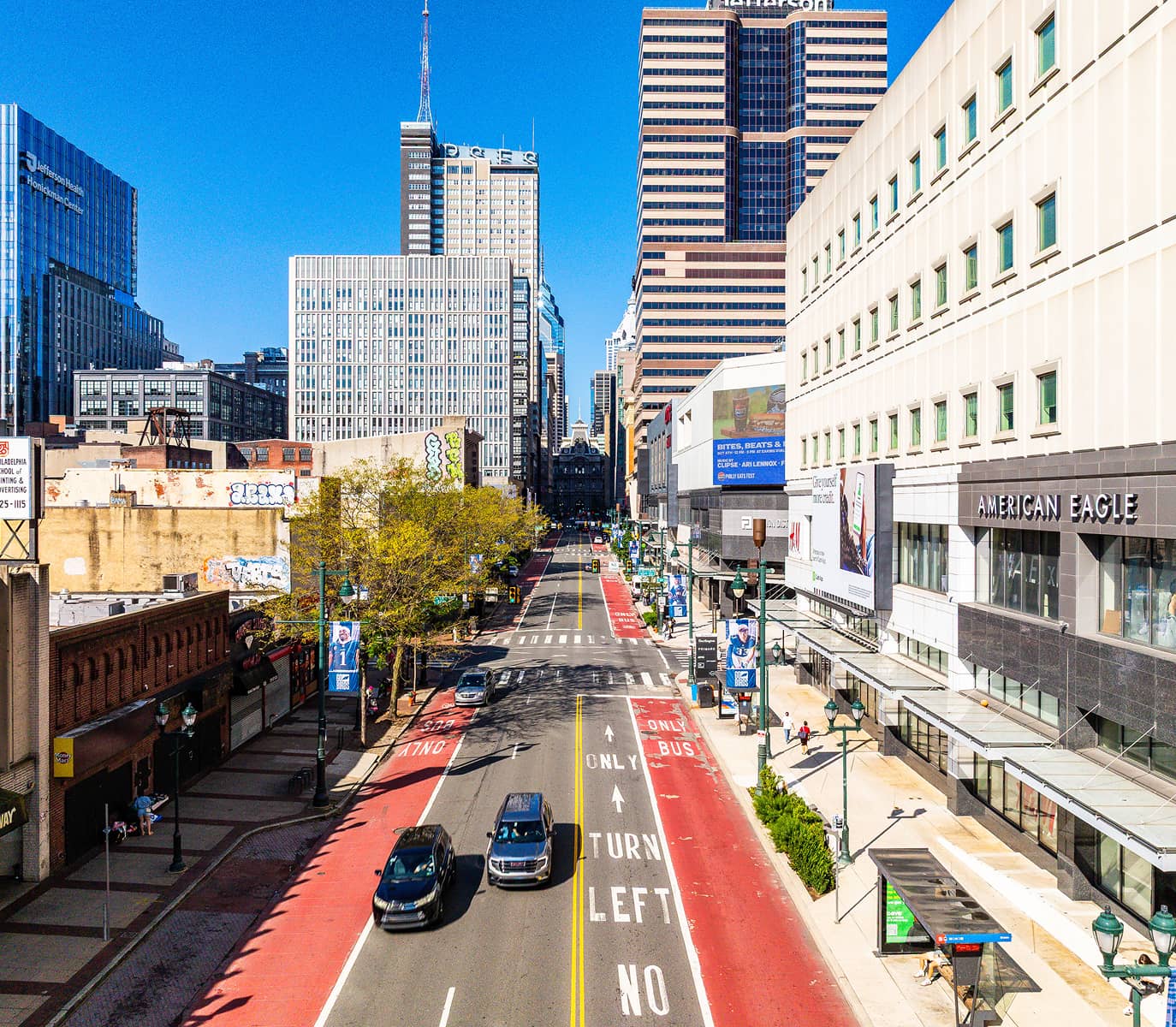 A vibrant city street with tall buildings and a clear blue sky. Cars drive along red bus lanes. Pedestrians walk by shops, including American Eagle.