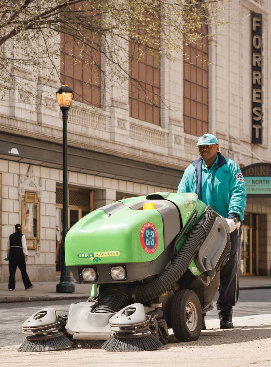 A street cleaner operates a bright green street-sweeping machine on a city sidewalk. The man wears a blue jacket and cap, with a theater and trees nearby.