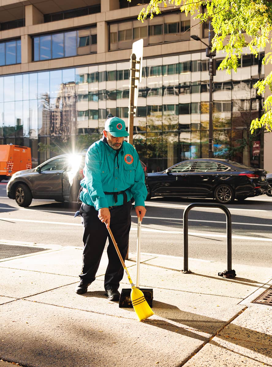 A city worker in a teal uniform sweeps the sidewalk in bright sunlight. Behind is a busy street with parked cars and a glass-fronted building. The scene feels industrious and orderly.