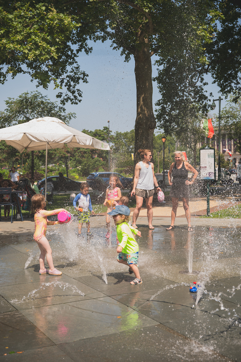 Toddlers playing in a public park with splash pad fountain and parents are in the background